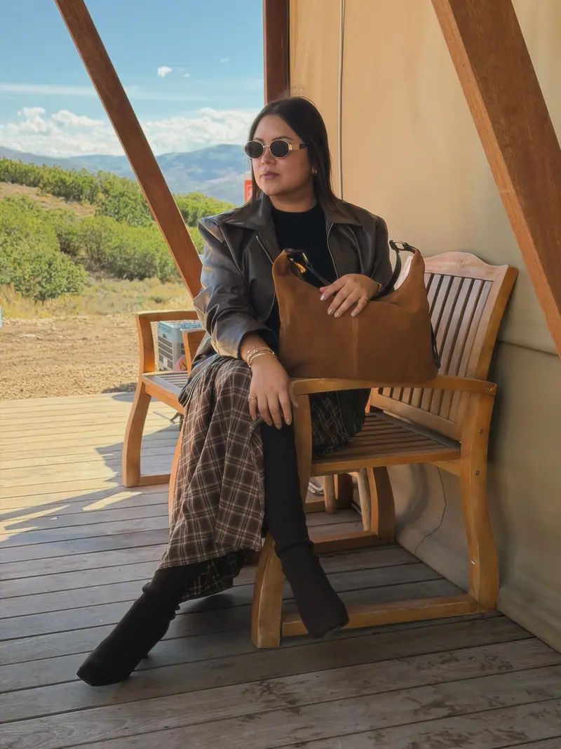 Seated on a wooden bench in a leather jacket with a caramel suede shoulder bag