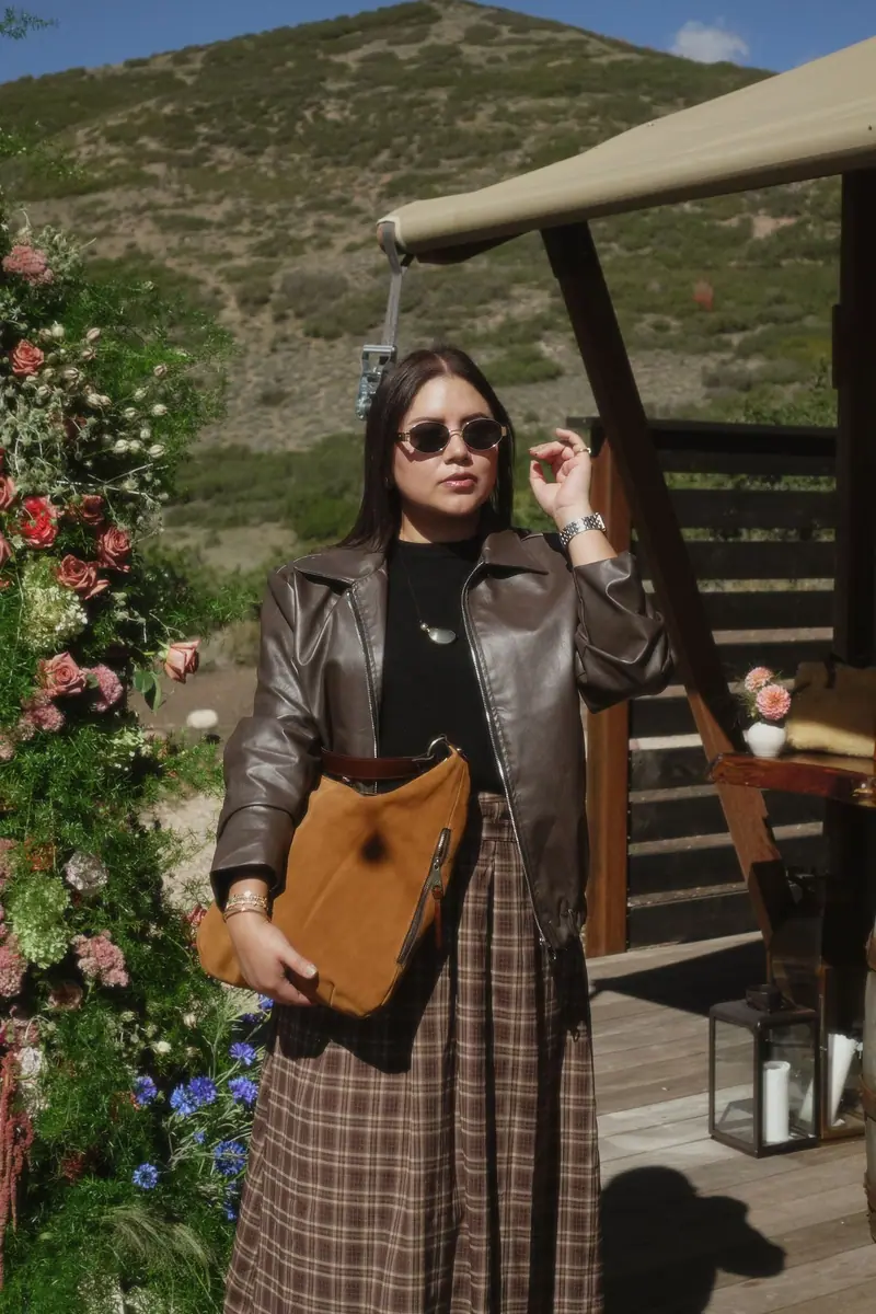 Brown leather jacket and sunglasses beside a rose and greenery arch at a mountain event